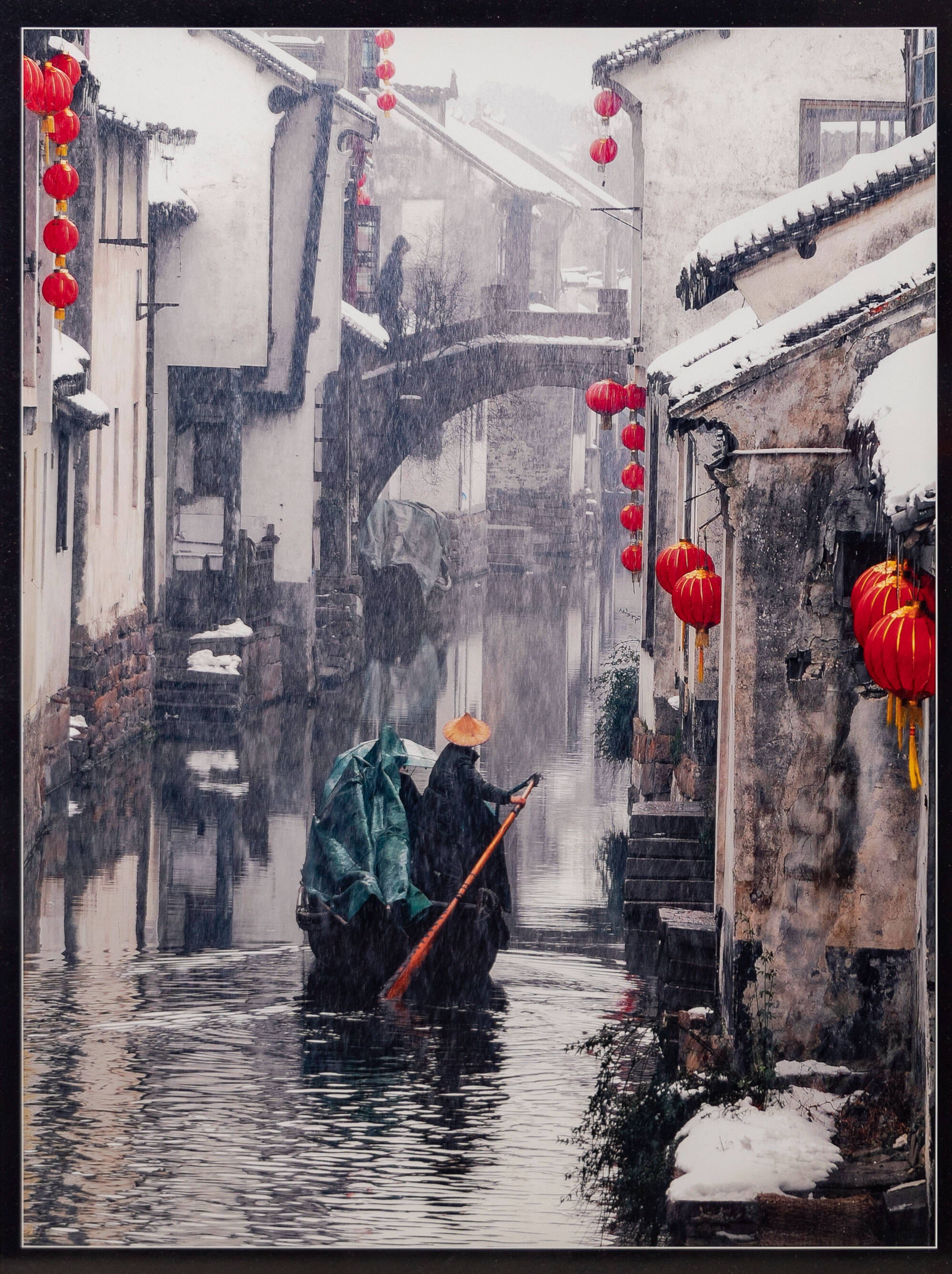 Snowy winter scene along a canal going through row of gray concrete houses. Subject is the back of a man in a boat going down canal.